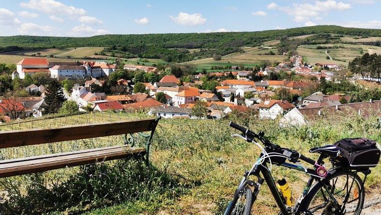 Radweg auf die Riede Hundschupfen, © Weinstraße Weinviertel Fahrrad und Bank mit Blick auf ein Dorf in hügeliger Landschaft.