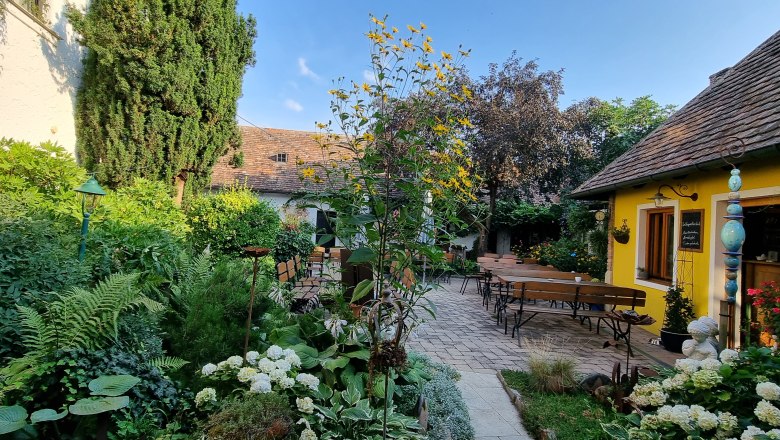 Blick in den Garten, © Fam. Lehninger Ein gemütlicher Garten mit gelbem Gebäude, Holzbänken und üppiger Vegetation unter blauem Himmel.