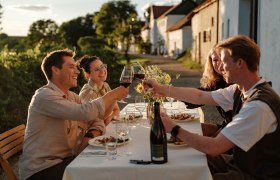 Weinviertler Kellergassenkulinarium, © Weinstraße Weinviertel / Reidinger Eine Gruppe von Menschen stößt bei einem Abendessen im Freien mit Weingläsern an. Im Hintergrund sind Weinberge und Häuser zu sehen.