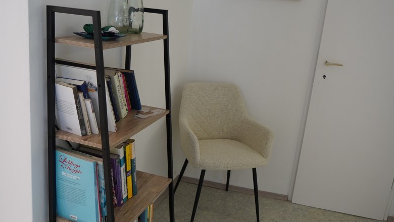 Landhaus Krell, © Weingut Krell Ein Bücherregal mit Büchern und Dekoration, daneben ein beiger Stuhl und eine Tür in einem Raum.