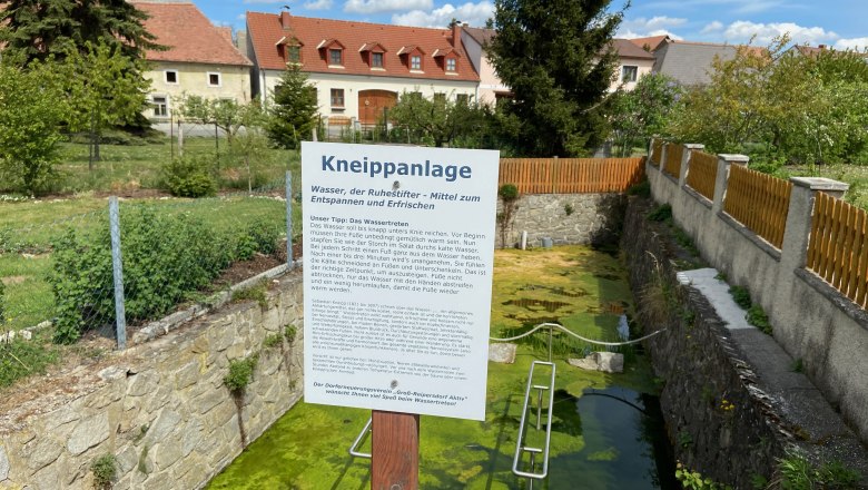 Wassertretanlage Großreipersdorf, © Weinstraße Weinviertel Kneippanlage mit Infotafel und Wasserbecken, umgeben von Zaun und Häusern im Hintergrund.