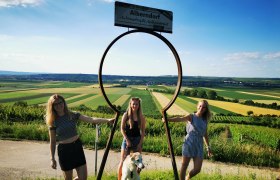Schlüsselerlebnis, © Weinstraße Weinviertel Drei Frauen und ein Hund posieren vor einem großen Schlüssellochrahmen mit Blick auf eine ländliche Landschaft.