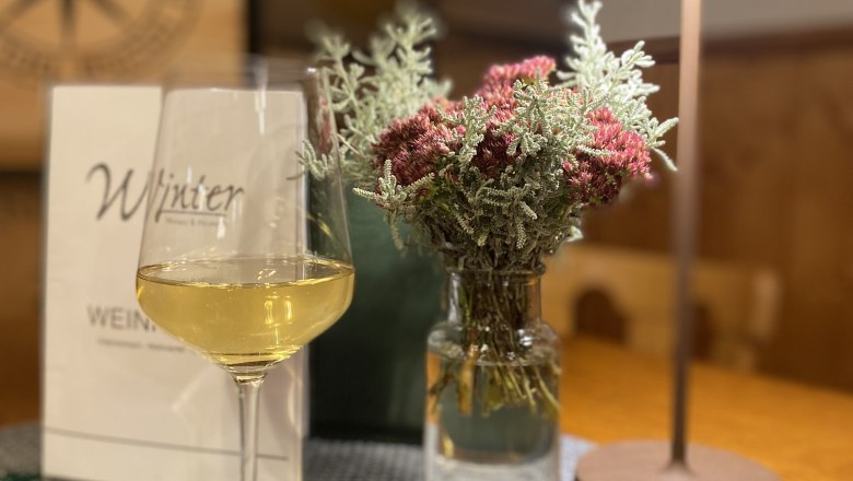 Weinbau und Heuriger Winter, © Cathy Winter Ein Weinglas mit Weißwein steht auf einem Tisch neben einer Vase mit Blumen und einer Tischlampe.