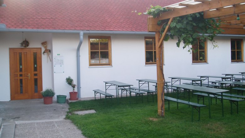 Garten, © Fam. Wallig Ein Garten mit Biertischen, einem Holzpergola und einem Gebäude mit roten Dachziegeln.