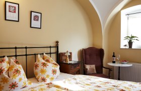 Angenehme Träume im Gästehaus Elisabeth, © Katharina F-Roßboth Gemütliches Gästezimmer mit Blumenbettwäsche, Sessel, Tisch mit Wein und Obst, und Pflanzen auf der Fensterbank.