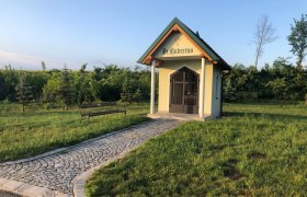 Hubertuskapelle Windpassing, © Petra-Eva Grüneis Kleine Kapelle mit Kreuz und Schriftzug "St. Hubertus" auf einer Wiese, mit gepflastertem Weg und Bank.