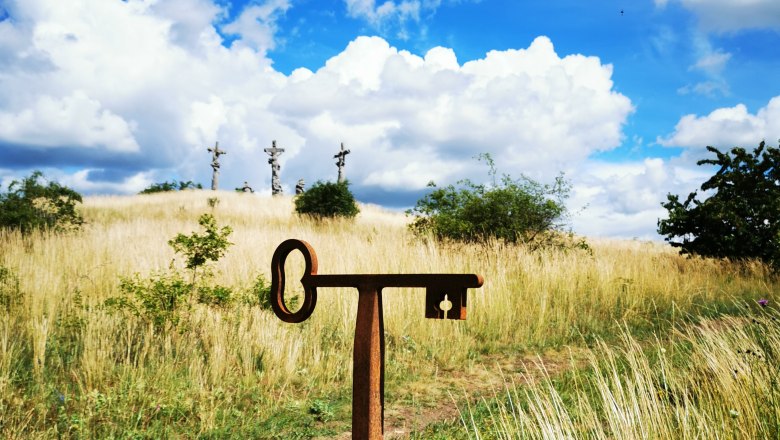 Perfekter Fotopunkt am Kalvarienberg, © Weinstraße Weinviertel Ein Fotopunkt-Schild vor einer Wiese mit drei Kreuzen im Hintergrund unter blauem Himmel mit Wolken.