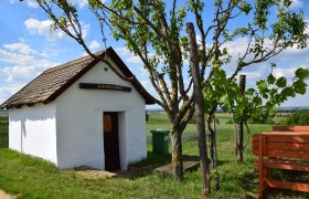 Hiatahittn Mittergrabern, © Christian Häusler Kleine weiße Hütte mit Holzdach und Schild "Hiatahitt'n Platzl". Daneben ein Baum und eine Bank, im Hintergrund Felder und blauer Himmel.