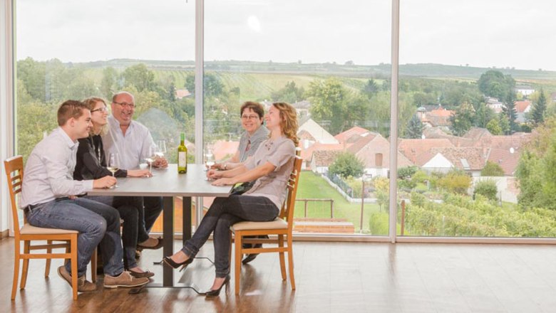 Familie Dworzak, © Astrid Bartl Eine Familie sitzt an einem Tisch mit Wein, im Hintergrund eine Landschaft mit Häusern und Feldern.