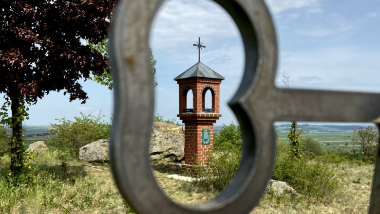 Herrgottsitz Etzmannsdorf, © Weinstraße Weinviertel Ein kleines Backsteinkapellchen mit Kreuz auf einem Hügel, durch ein herzförmiges Metallgitter fotografiert.