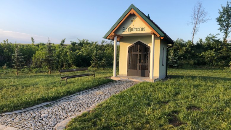 Hubertuskapelle Windpassing, © Petra-Eva Grüneis Kleine Kapelle mit Kreuz und Schriftzug "St. Hubertus" auf einer Wiese, mit gepflastertem Weg und Bank.
