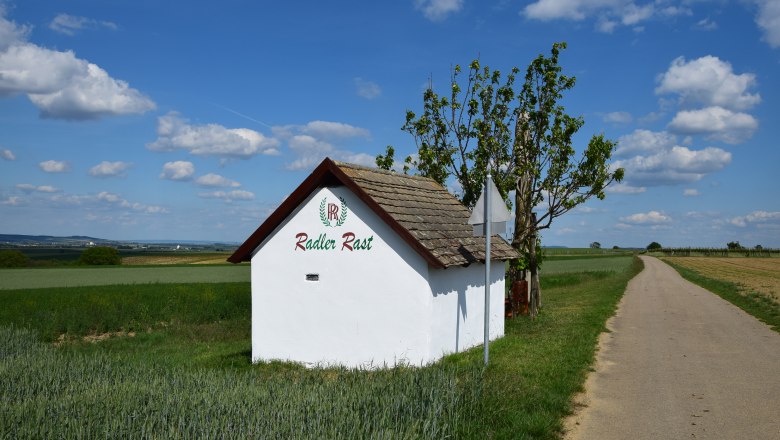 Hiatahittn Mittergrabern, © Christian Häusler Kleines weißes Gebäude mit der Aufschrift 'Radler Rast' neben einem Feldweg unter blauem Himmel.