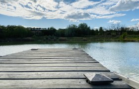 Idyllisches Fischerparadies, © Weinstraße Weinviertel Holzsteg am Ziegelofenteich in Zellerndorf, umgeben von Bäumen und blauem Himmel mit Wolken.