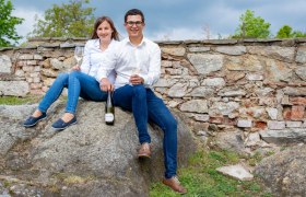 Weingut und Buschenschank Freytag, © Martin Lifka Photography Ein Mann und eine Frau sitzen auf einem Felsen vor einer Steinmauer, halten Weingläser und eine Weinflasche.