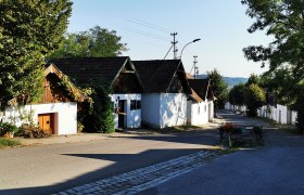 Kellergasse Lumpenspitz, © Weinstraße Weinviertel Kellergasse mit weißen Häusern und Bäumen bei Sonnenschein.