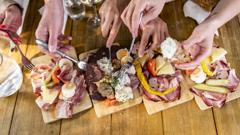 Heurigenjause, © Weinviertel Tourismus GmbH / Herbst Mehrere Hände greifen nach Wurst- und Käseplatten auf einem Holztisch.