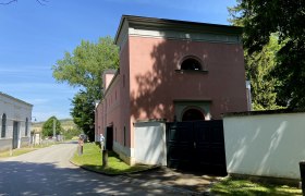 Heldenberg beim Schloss, © Weinstraße Weinviertel Straße mit rosa Gebäude und Bäumen bei sonnigem Wetter.