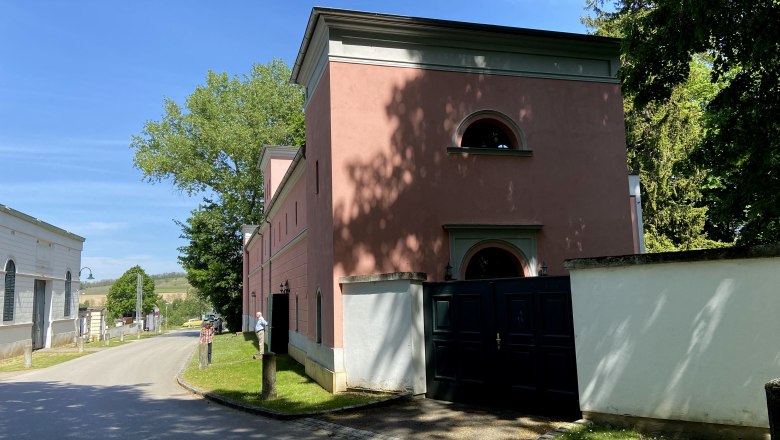 Heldenberg beim Schloss, © Weinstraße Weinviertel Straße mit rosa Gebäude und Bäumen bei sonnigem Wetter.