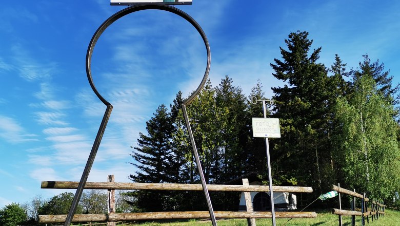 Hubertuskapelle Alberndorf, © Weinstraße Weinviertel Großer Schlüssellochrahmen mit Schild 'Alberndorf' und 'Foto Punkt' vor Bäumen und blauem Himmel.