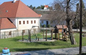Kinderspielplatz beim Pfarrhof, © Souveräner Malteser-Ritter-Orden / Udo Schwamberger Ein Kinderspielplatz mit Klettergerüst und Schaukel neben einem Gebäude mit rotem Dach.