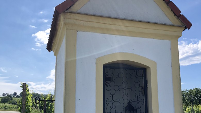 Weinbergkapelle von Glaubendorf, © Heldenberg Weinbergkapelle von Glaubendorf mit schmiedeeisernem Tor und Schild 'Geheimtipp' daneben, umgeben von grüner Landschaft.