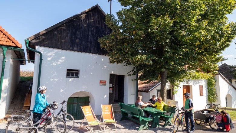 Keller-Wohlführplatz Alberndorf, © Initiative Pulkautal /P. Mödl Menschen sitzen auf Bänken vor einem weißen Gebäude mit Fahrrädern in Alberndorf.