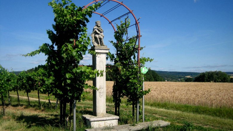 Mautkreuz Ronthal, © Marktgemeinde Hohenwarth-Mühlbach Steinernes Kreuz mit Figur in ländlicher Umgebung, umgeben von Pflanzen und Feldern.