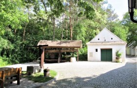 Weinvierlter Kellergassenkulinarium, © D`Raschalaer Köllamauna Eine gepflasterte Kellergasse mit einem kleinen weißen Gebäude und einer überdachten Holzpresse, umgeben von Bäumen.