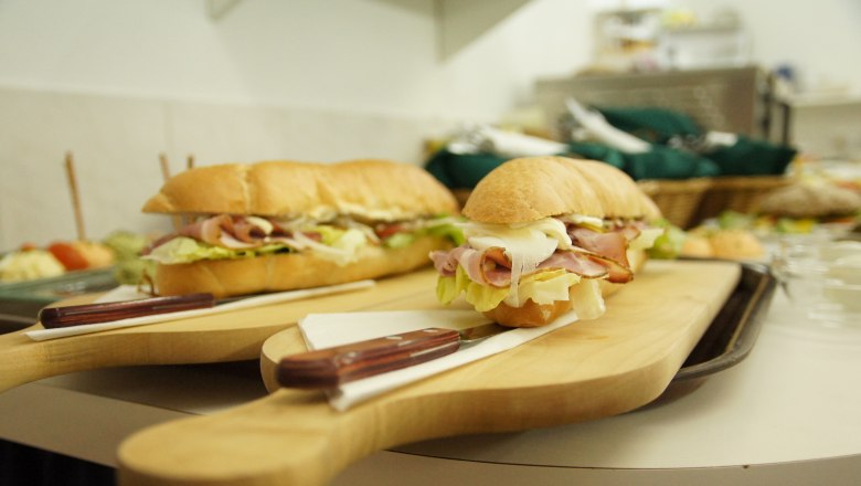 Heurigenjause, © Fam. Zöchmeister Zwei belegte Baguettes auf Holzbrett mit Messer, im Hintergrund weitere Speisen.