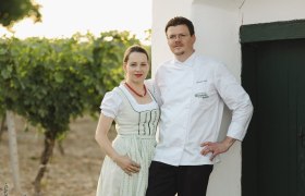 Wirtsleute Daniela und Harald Graf, © Niederösterreich Werbung/Michael Reidinger Ein Paar in traditioneller Kleidung steht vor einem weißen Gebäude neben einem Weinberg.