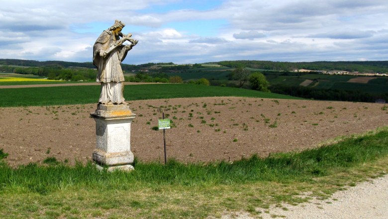 Nepomuk Statue in Baierdorf, © Marktgemeinde Ravelsbach Statue von Nepomuk auf einem Feld in Baierdorf, umgeben von grüner Landschaft und bewölktem Himmel.