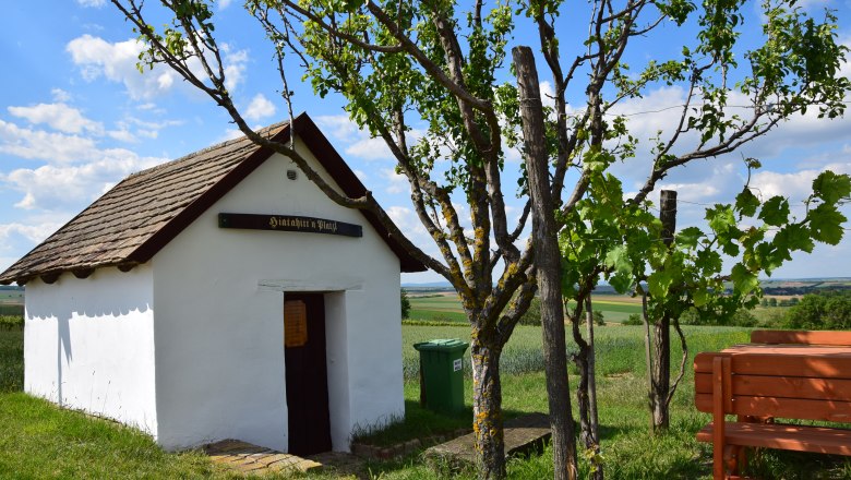 Hiatahittn Mittergrabern, © Christian Häusler Kleine weiße Hütte mit Holzdach und Schild "Hiatahitt'n Platzl". Daneben ein Baum und eine Bank, im Hintergrund Felder und blauer Himmel.