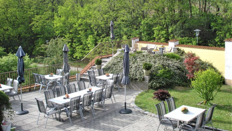 Gästeterrasse, © Pension Rudi Gästeterrasse mit Tischen, Stühlen und Sonnenschirmen, umgeben von grüner Vegetation.