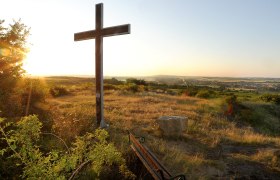 Sonnenuntergang am Altenberg, © H. Schleich Ein Kreuz auf einem Hügel bei Sonnenuntergang mit Blick auf eine weite Landschaft und ein Dorf im Hintergrund.
