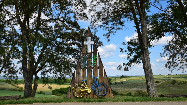 Radfahrer Marterl in Mittergrabern, © Christian Häusler Ein Marterl mit einem gelben und blauen Fahrrad, umgeben von Bäumen und Feldern unter blauem Himmel.