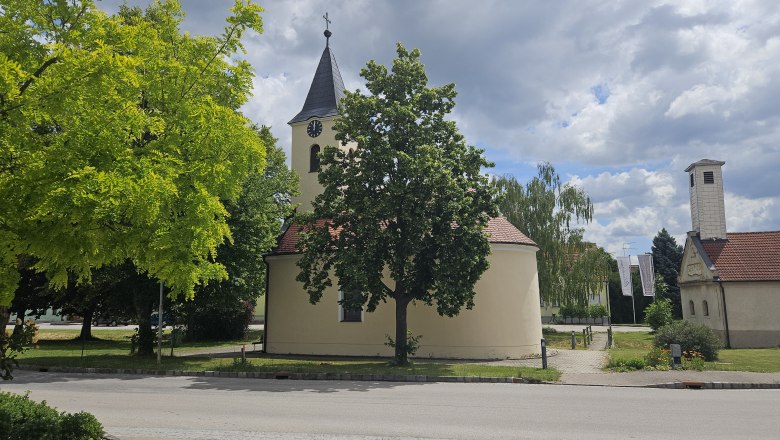 Barbara Kapelle Kleinweikersdorf, © Barbara Sturmlechner Eine kleine Kapelle mit einem Turm und einer Uhr, umgeben von Bäumen und einem blauen Himmel mit Wolken.