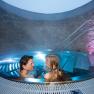 Vino, © Althof Retz Ein Paar entspannt in einem runden Whirlpool mit blauer Beleuchtung und Steinwänden.