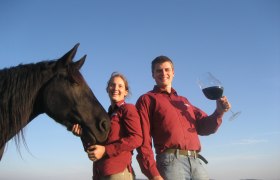 weinkutscher.at, © Stefan Reinthaler Zwei Personen in roten Hemden mit einem schwarzen Pferd und einem großen Weinglas im Freien.