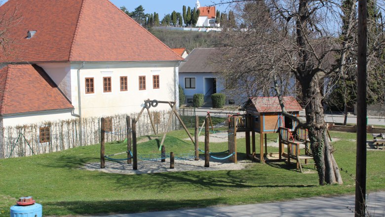 Kinderspielplatz beim Pfarrhof, © Souveräner Malteser-Ritter-Orden / Udo Schwamberger Ein Kinderspielplatz mit Klettergerüst und Schaukel neben einem Gebäude mit rotem Dach.