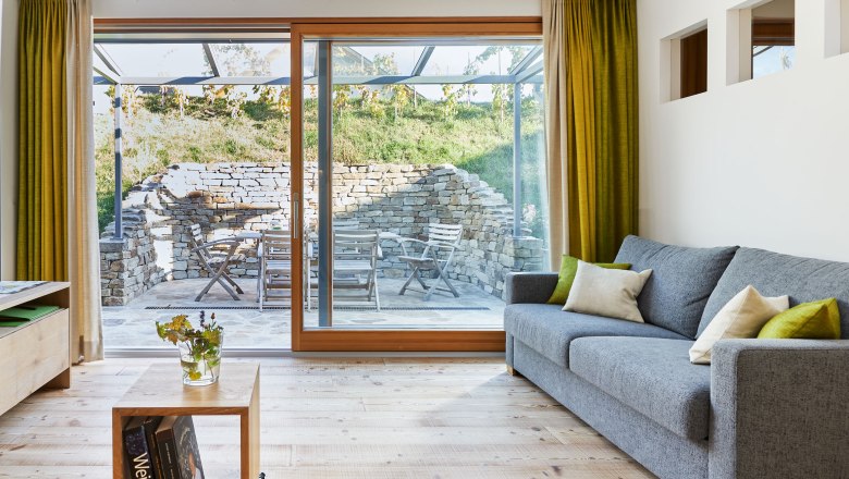 Urlaub im Weingut Hindler, © Andreas Hofer Modernes Wohnzimmer mit grauem Sofa, Holzboden und großen Fenstern mit Blick auf eine Terrasse mit Steinmauer und Gartenmöbeln.