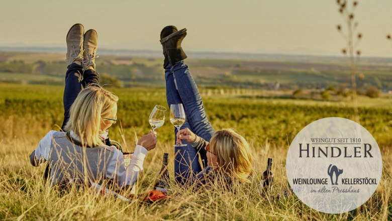 Zu Gast im Weingut Hindler, © Weingut Hindler Zwei Personen liegen in einem Weinberg, heben die Füße und halten Weingläser. Logo des Weinguts Hindler ist eingeblendet.