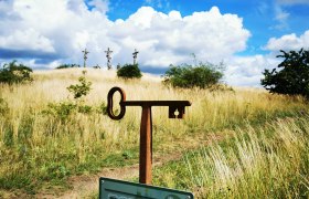 Kalvarienberg Schrattenthal, © Weinstraße Weinviertel Ein Fotopunkt-Schild vor einer Wiese mit drei Kreuzen im Hintergrund unter blauem Himmel mit Wolken.