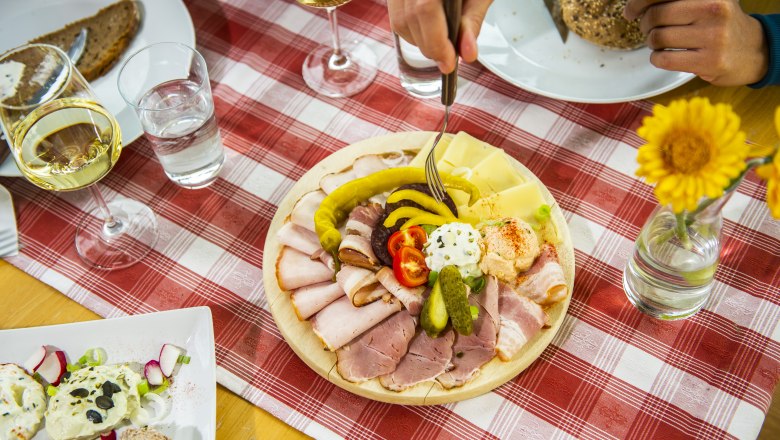 Heurigenjause, © POV / Robert Herbst Ein gedeckter Tisch mit einer Platte voller Aufschnitt, Käse und Gemüse, daneben Gläser mit Weißwein und Wasser auf einer rot-weiß karierten Tischdecke.
