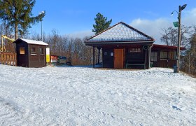 Schneebedeckte Schiclub-H&uuml;tte mit Holzh&auml;uschen und B&auml;umen im Hintergrund.