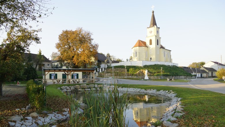 Dorfplatz in Glaubendorf mit Kirche, Teich und Herbstbäumen.