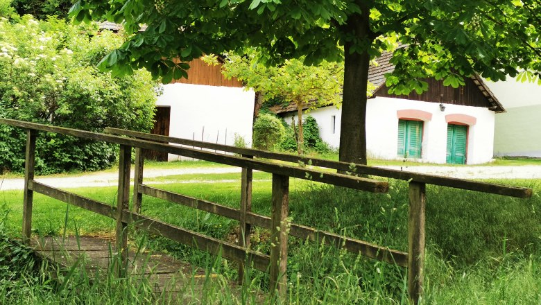 Eine kleine Holzbrücke führt über eine Wiese zu weißen Gebäuden mit grünen Türen, umgeben von Bäumen und Büschen.