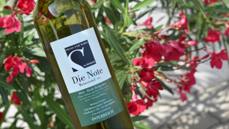 Weinflasche mit Etikett "Die Note" vor blühenden roten Blumen.