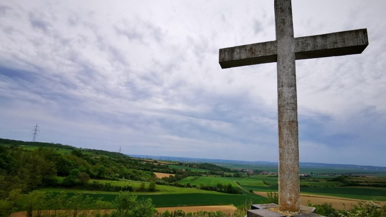 Ein großes Steinkreuz auf einem Hügel mit Blick auf eine grüne Landschaft und bewölkten Himmel.