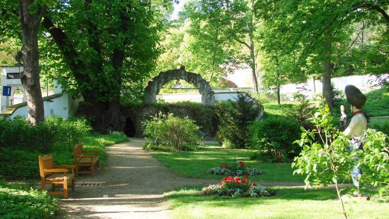 Ein grüner Garten mit Bäumen, Bänken und einer Statue.