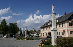 Ein Platz in Weitersfeld mit einer Statue, mehreren Flaggen und einem Gebäude im Hintergrund.